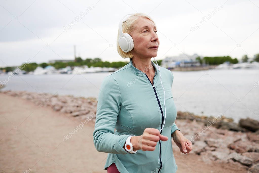 Mature female with headphones listening to music or audio while jogging along riverside in urban environment in the morning