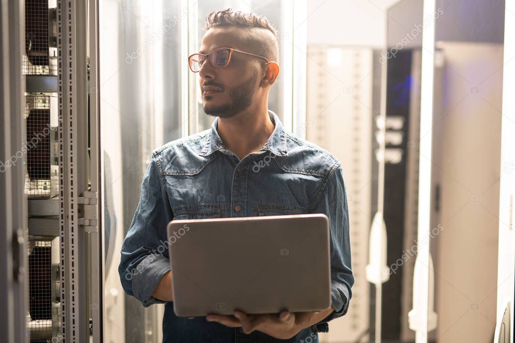 Serious confident young Arabian IT engineer with beard standing in data center and using laptop while providing safety of network server