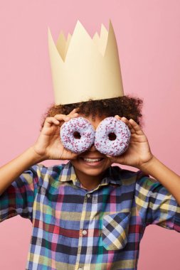Waist up portrait of smiling African-American boy holding donuts posing against pink background, Birthday party concept clipart