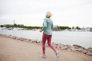 Sabah ları su kenarında koşarken aktif giyimli sarışın sporcunun arka dansı kulaklıkla müzik dinliyor