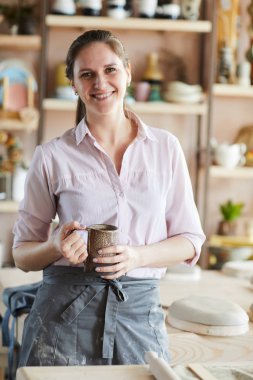 Waist up portrait of cheerful female artisan posing in pottery studio holding handmade mug, copy space clipart