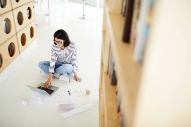 Konsantre kadın düz tasarımcı planı ve buruşturulmuş kağıtları ile katta oturan ve kütüphanede kroki üzerinde çalışan yukarıda görünümü