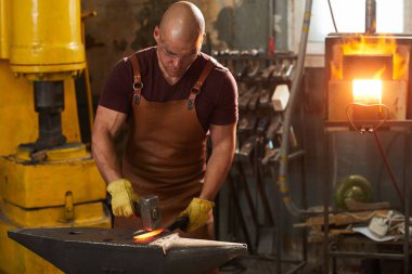 Deri önlük ve koruyucu eldiven odaklanmış genç adam örs ayakta ve çekiç ile sıcak demir dayak