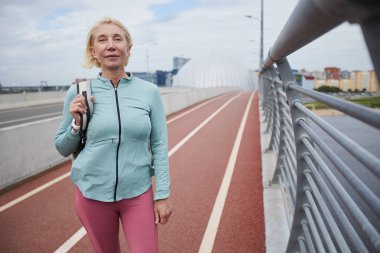 Boş zamanlarında kamera önünde açık stadyumda yarış pistinde duran sırt çantası ile Aktif olgun sarışın kadın