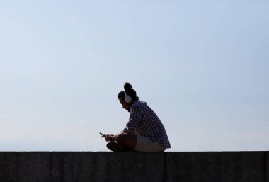 Mavi gökyüzüne karşı ana hatlarıyla beton blok üzerinde otururken müzik dinlerken plajda ürpertici çağdaş genç adamın yan görünüm portresi, kopya alan