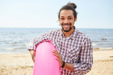 Pembe yüzme tüpü, eğlenceli Yaz konsepti tutan plajda poz verirken kamera önünde mutlu bir şekilde gülümseyen genç Latin-Amerikalı bir adamın portresi