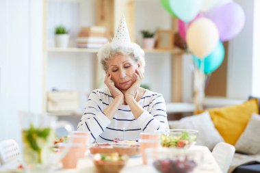 Parti şapkası ciddi mutsuz emekli gri saçlı bayan yemek masasında yemek masası tabaklarla dolu oturan ve yalnız doğum günü partisi yaparken mum ile doğum günü pastası bakarak.