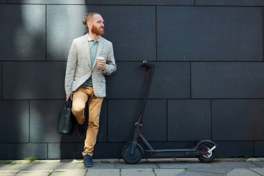 Sakallı pozitif aktif hipster genç işadamı sokakta bina duvara yaslanmış ve kahve içme, gidip için modern kick scooter