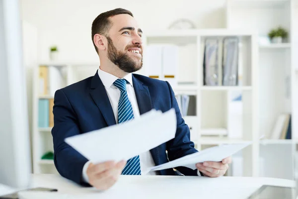 Positive excited young bearded financial manager in suit sitting at table in office and analyzing business documents