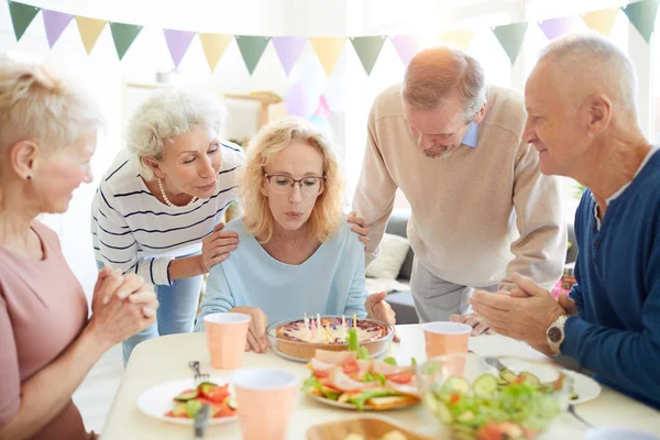 Beautiful mature lady in glasses supported by friends blowing candles at birthday party, copy space