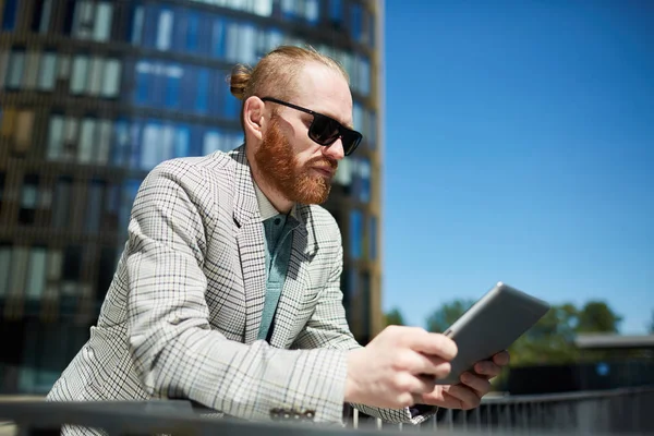 Serious thoughtful young bearded manager in sunglasses leaning on railing in city and viewing information on tablet