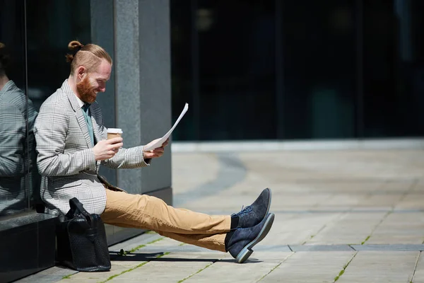 Positive successful hipster young lawyer with beard sitting outdoors and reading papers while preparing for court pleading