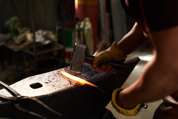 Close-up of unrecognizable blacksmith standing at steel anvil and shaping heated metal bar with hammer in workshop