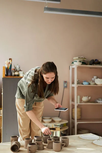 Portrait of female artisan taking smartphone pictures of handmade ceramic items in pottery shop, copy space