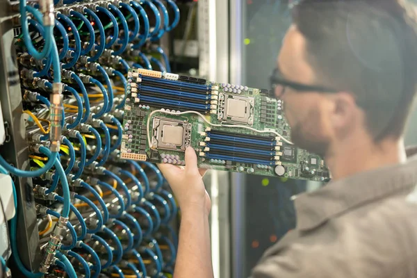 Busy skilled young IT expert in glasses standing by server cabinet and analyzing motherboard of supercomputer