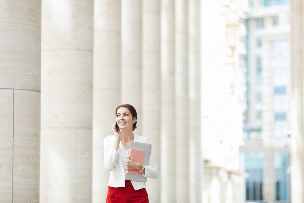 Waist up portrait of beautiful woman speaking by phone while walking towards camera along row of pillars, copy space