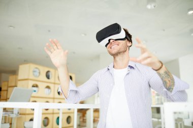 Neşeli modern kütüphanede ayakta rahat gömlek giyen ve video oyunu oynarken sanal gerçeklik simülatörü kullanarak stubble ile neşeli dövmeli adam