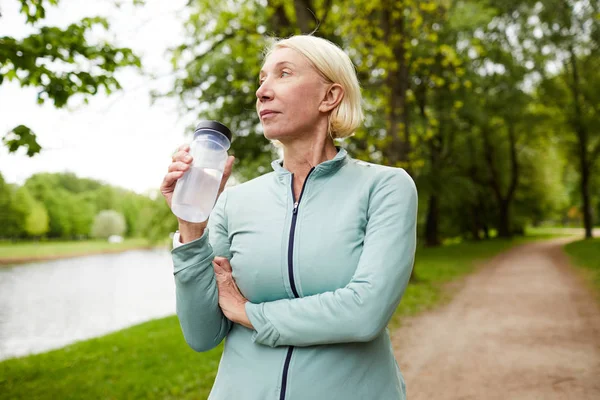Doğal ortamda egzersiz sonrası dinlenme sırasında aktif giyim su plastik şişe tutan sakin olgun kadın