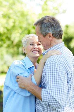 Waist up portrait of happy senior couple looking at each other while embracing in Summer park clipart