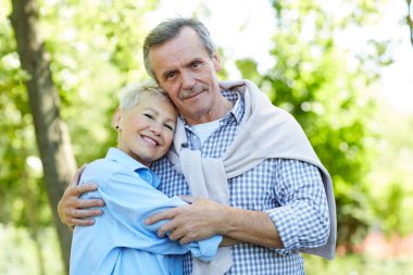 Waist up portrait of happy senior couple embracing and looking at camera while enjoying walk in Summer park, copy space clipart