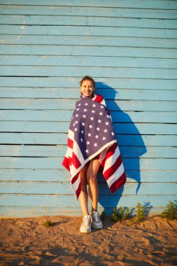 İçerik genç kadın mavi bina cephe karşı kum üzerinde duran ve kameraya bakarak Amerikan bayrağı sarılı