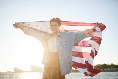 Abd'de ulusal günü kutlarken sahilde duran ve Amerikan bayrağı tutan açık kot ceketli neşeli heyecanlı genç kadının portresi