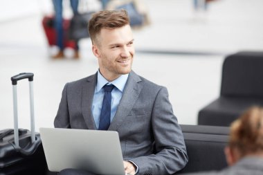 Havaalanında bagajla oturan ve toplantı hazırlığı nda dizüstü bilgisayar kullanan takım elbiseli başarılı bayi