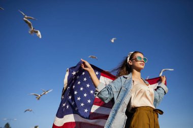 Güneş gözlüğü ve başörtüsü ciddi şık kız görünümü aşağıda rüzgar ayakta ve mesafe bakarken Amerikan bayrağı afiş tutan, havada martılar