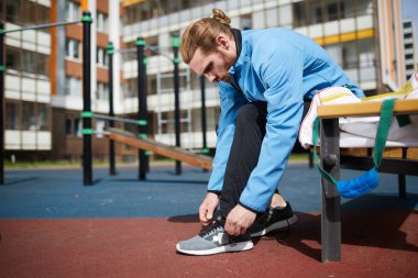 At kuyruğu olan ciddi yakışıklı genç adam ceket ve spor pantolon giyen açık zemin de bankta oturan ve spor ayakkabı dantel bağlama