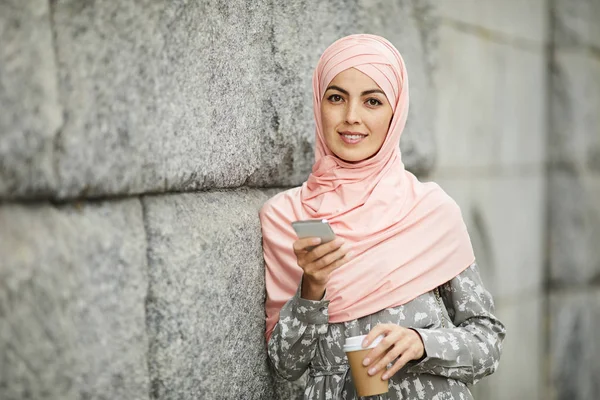 积极的现代阿拉伯商务女士与外带咖啡杯靠在墙上和在电话检查笔记 — 图库照片