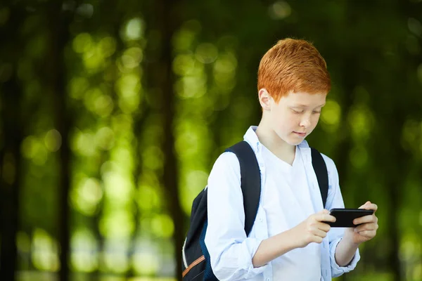Redheaded schoolboy playing with his phone on his way home in park after classes at school