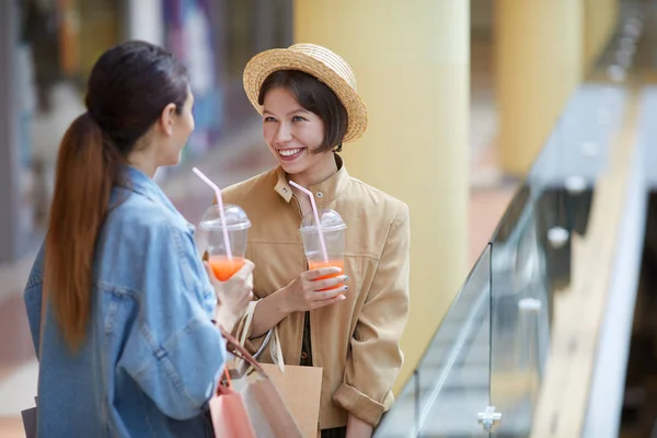 Happy excited talkative female shoppers in casual outfits standing at railing in mall and enjoying sweet drinks