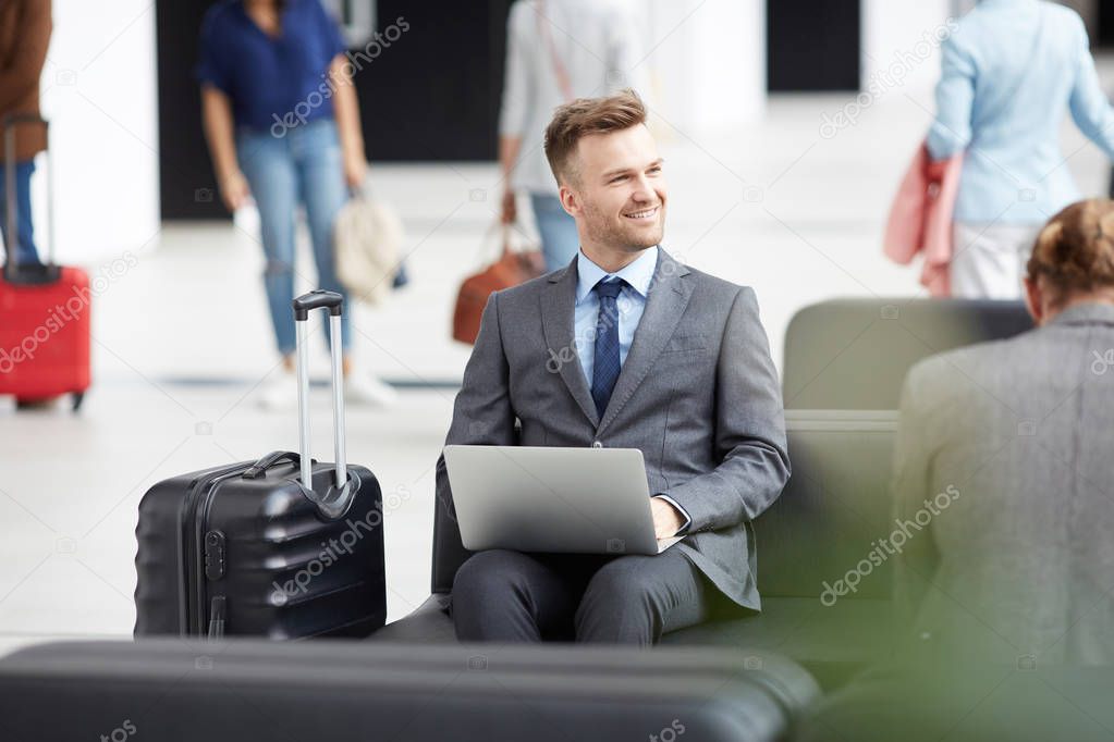 Positive inspired young entrepreneur in formalwear sitting in airport area using laptop while working on presentation