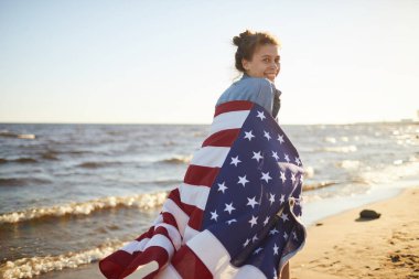 Amerikan bayrağı ile sahil boyunca çalışan Amerikan bayrağı sarılı olumlu çekici genç kadın Arka görünümü