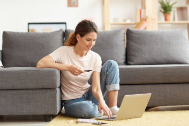 Çekici genç bir kadın evde halıda oturuyor ve internette alışveriş yaparken dizüstü bilgisayar kullanıyor.