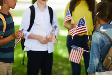 Bir grup modern Amerikalı çocuk, dışarıda daire çizip Amerikan kültürü hakkında konuşurken bayraklar dikiliyor.