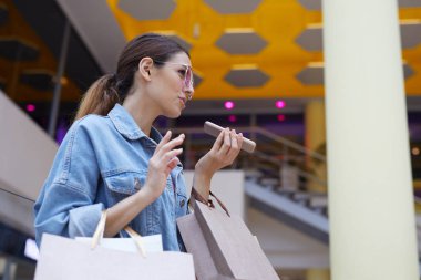 Mavi kot ceketli genç kız büyük yeni alışveriş merkezinin ortasında duran arkadaşı için sesli mesaj yapma
