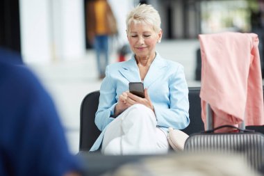 Mavi ceketli gülümseyen sarışın kadın havaalanında deri koltukta oturan ve gadget wifi kullanarak