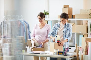 İki genç kadın online müşteriler için parsel ambalaj - kutulara şeyler koyarak ve viyolonsel ile mühürleme