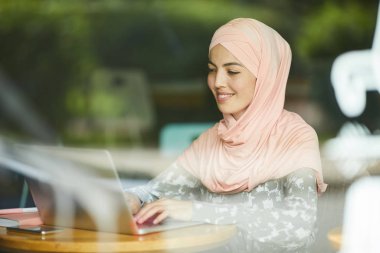 Laptoptaki e-maillere cevap veren pembe tesettürlü oldukça olumlu bir genç kadın.