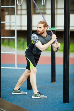 Spor giyimli, dış mekanda duran ve eforla halatı çeken, ifade veren genç sakallı adam kaşlarını çatarak