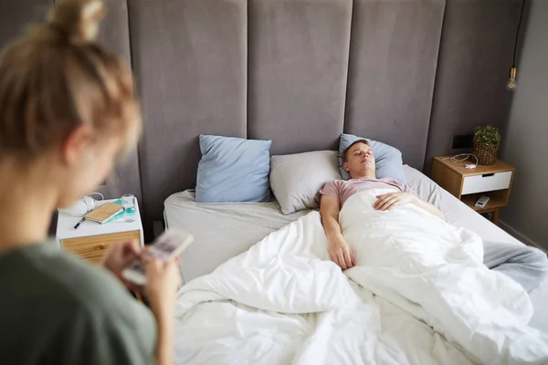 Young tired man sleeping in bed while wife or girlfriend scrolling in his smartphone near by