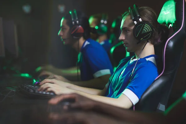 Group of concentrated video game club participants in modern headsets sitting in row and playing computer game