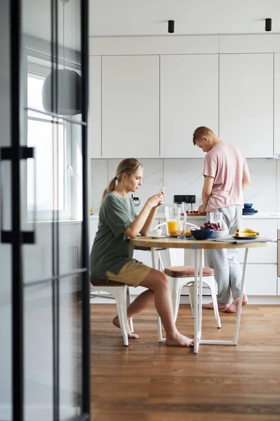 台所のテーブルのそばに座り 朝食を食べ 夫が料理をしながらスマートフォンを使う若い女性 — ストック写真