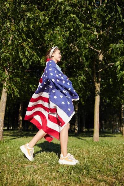stock image Smiling excited pretty girl wearing headscarf and stylish sneakers wrapped in big American flag walking in summer park