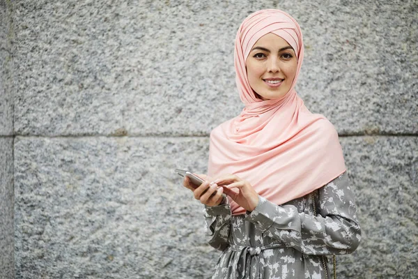 正积极的年轻阿拉伯妇女在粉红色头巾站在老建筑和使用智能手机上的移动互联网的肖像 — 图库照片