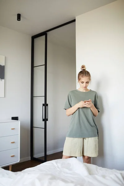 寝室のベッドに立っている間 かなり若いカジュアルな女性がスマートフォンでスクロール — ストック写真