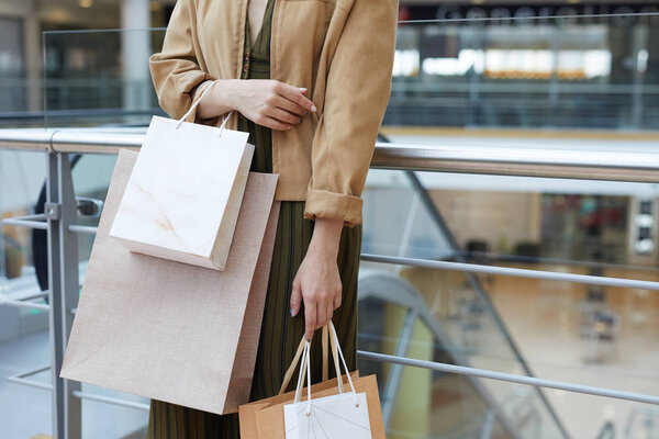 Close-up of stylish lady in jacket and dress standing at railing in mall and holding shopping bags