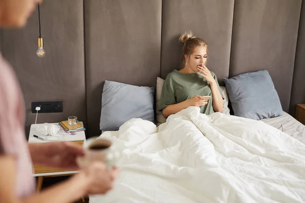 彼女の夫が彼女のコーヒーを持って来ている間 朝ベッドに座ってスマートフォンを持つ若いあくびの女性 — ストック写真