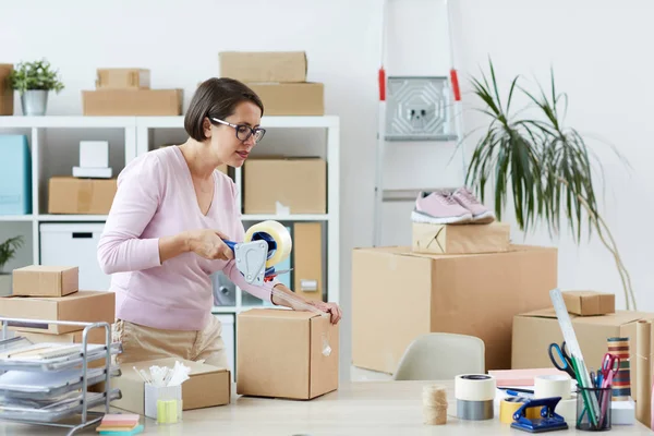 Young manager in casualwear packing box with online order of client with cellotape in office